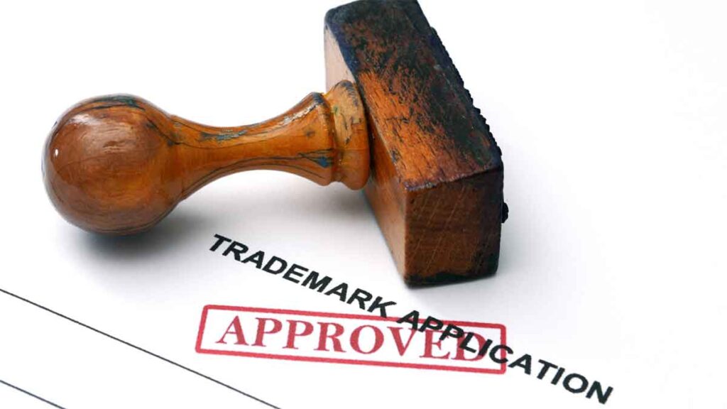 A wooden stamp on a paper labeled _Trademark Application_ with a red _APPROVED_ stamp mark