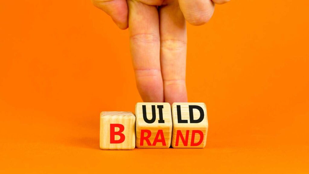 A hand arranging wooden blocks spelling _BUILD BRAND_ on an orange background